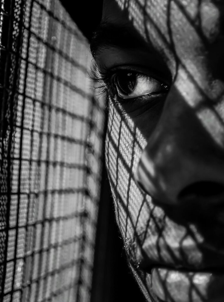 Por Que Aceitamos Migalhas Emocionais? 
Foto em preto e branco de um close no olho de uma pessoa, que aparece parcialmente coberto por sombras projetadas por uma grade ou tela. O olhar transmite tristeza e aprisionamento emocional, simbolizando a sensação de estar preso a migalhas afetivas e relações que não oferecem o que realmente se merece.