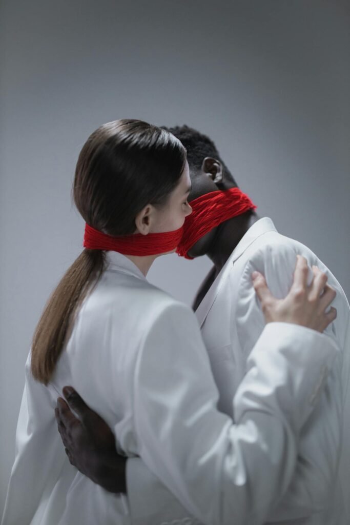 Imagem conceitual de um casal vestido de branco, com pano vermelho envolvendo suas bocas, simbolizando silêncio e aprisionamento emocional. Representa a falta de voz e a perda de autonomia em relacionamentos abusivos com pessoas narcisistas.