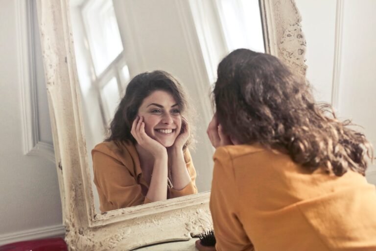 Mulher sorrindo para si mesma no espelho, representando o fortalecimento da autoestima e do amor-próprio.