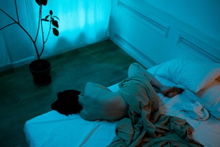 Mulher deitada na cama em um quarto escuro, cercada por luz azul, representando um momento de solidão e introspecção.